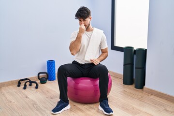 Hispanic man with beard sitting on pilate balls at yoga room smelling something stinky and...