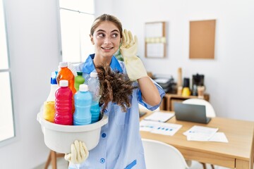 Young blonde woman wearing cleaner uniform holding cleaning products smiling with hand over ear...