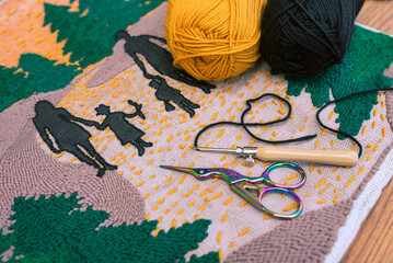 Small scissors, punch needle and two skeins of yarn on embroidered picture of family walking in the...