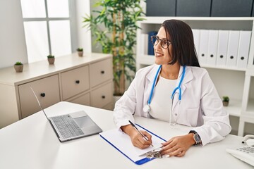 Young hispanic woman wearing doctor uniform writing on clipboard at clinic