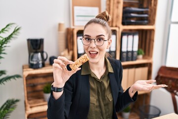 Young hispanic girl eating protein bar as healthy energy snack at the office celebrating achievement with happy smile and winner expression with raised hand