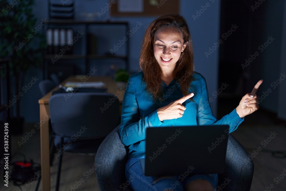 Poster brunette woman working at the office at night smiling and looking at the camera pointing with two ha