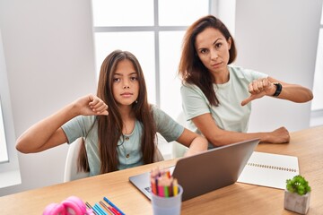 Young mother and daughter doing homework at home with angry face, negative sign showing dislike with thumbs down, rejection concept
