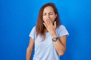 Brunette woman standing over blue background bored yawning tired covering mouth with hand. restless and sleepiness.