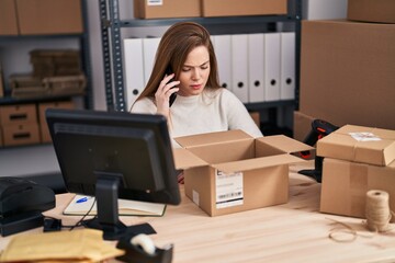 Fototapeta na wymiar Young blonde woman ecommerce business worker unboxing package and talking on the smartphone at office