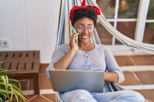 African american woman using laptop and talking on smartphone lying on hammock at home terrace