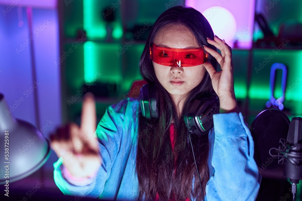 Poster Young chinese woman streamer playing video game using virtual reality glasses at gaming room