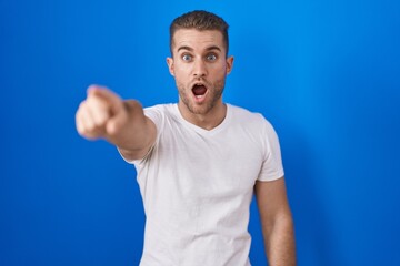 Naklejka premium Young caucasian man standing over blue background pointing with finger surprised ahead, open mouth amazed expression, something on the front