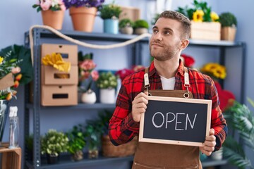 Young caucasian man working at florist holding open sign smiling looking to the side and staring away thinking.