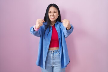 Young asian woman standing over pink background angry and mad raising fists frustrated and furious while shouting with anger. rage and aggressive concept.