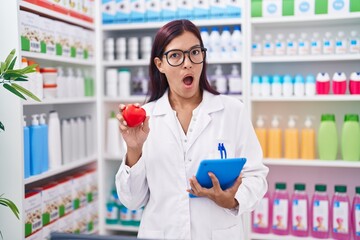 Young hispanic woman working at pharmacy drugstore holding red heart in shock face, looking skeptical and sarcastic, surprised with open mouth