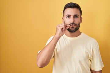 Handsome hispanic man standing over yellow background mouth and lips shut as zip with fingers....