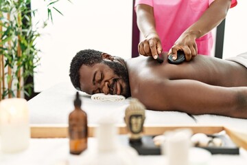 Obraz na płótnie Canvas African american man reciving back massage with black stones at beauty center.