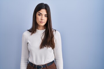 Obraz premium Young brunette woman standing over blue background relaxed with serious expression on face. simple and natural looking at the camera.