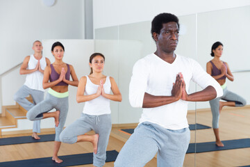 Obraz premium Focused aframerican man standing on one leg in Eka Pada Pranamasana pose during group yoga training in gym