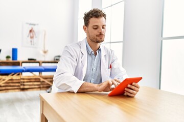 Young hispanic man wearing physiotherapist uniform using touchpad at clinic