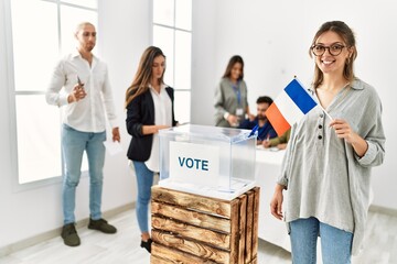 Young french voter woman smiling happy holding france flag standing by ballot at vote center.