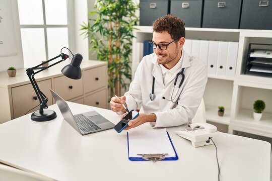 Young Hispanic Man Wearing Doctor Uniform Having Telemedicine Showing Smartphone At Clinic