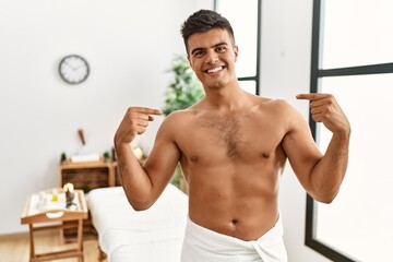 Obraz premium Young hispanic man standing shirtless at spa center looking confident with smile on face, pointing oneself with fingers proud and happy.