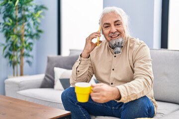 Fototapeta premium Middle age grey-haired man talking on smartphone drinking coffee at home