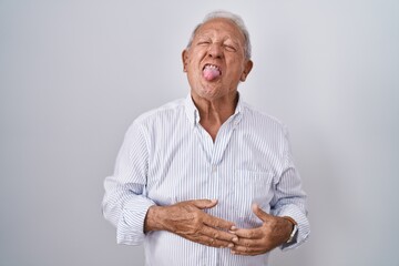 Senior man with grey hair standing over isolated background sticking tongue out happy with funny...