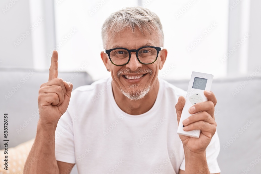 Sticker Hispanic man with grey hair holding air conditioner control smiling with an idea or question pointing finger with happy face, number one