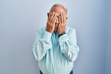 Senior man with grey hair standing over blue background with sad expression covering face with...