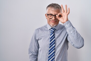 Hispanic business man with grey hair wearing glasses doing ok gesture with hand smiling, eye looking through fingers with happy face.