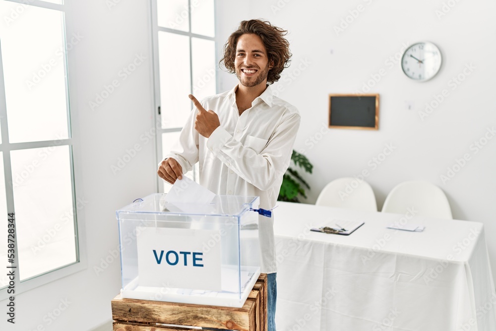 Poster Young hispanic man voting putting envelop in ballot box cheerful with a smile on face pointing with hand and finger up to the side with happy and natural expression