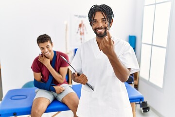 Obraz premium Young hispanic man working at pain recovery clinic with a man with broken arm beckoning come here gesture with hand inviting welcoming happy and smiling