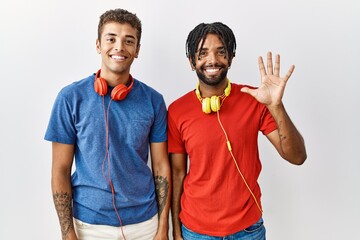 Young hispanic brothers standing over isolated background wearing headphones showing and pointing up with fingers number five while smiling confident and happy.