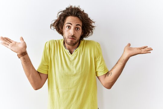 Young hispanic man standing over isolated background clueless and confused expression with arms and hands raised. doubt concept.