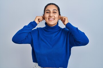Young hispanic woman standing over isolated background smiling pulling ears with fingers, funny...
