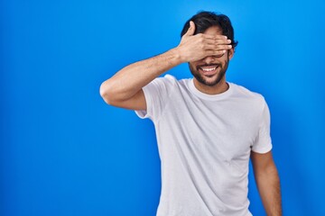 Handsome latin man standing over blue background smiling and laughing with hand on face covering eyes for surprise. blind concept.