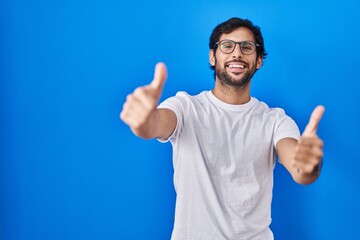 Handsome latin man standing over blue background approving doing positive gesture with hand, thumbs...