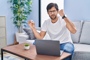 Handsome latin man holding virtual currency bitcoin using laptop annoyed and frustrated shouting with anger, yelling crazy with anger and hand raised