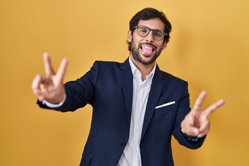 Handsome latin man standing over yellow background smiling with tongue out showing fingers of both...