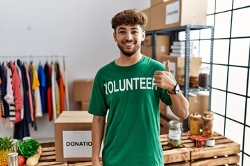 Young arab man wearing volunteer t shirt at donations stand doing happy thumbs up gesture with hand. approving expression looking at the camera showing success.