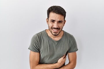 Young hispanic man with beard wearing casual t shirt over white background with hand on stomach because nausea, painful disease feeling unwell. ache concept.