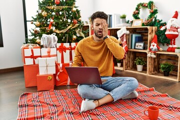 Arab young man using laptop sitting by christmas tree yawning tired covering half face, eye and mouth with hand. face hurts in pain.