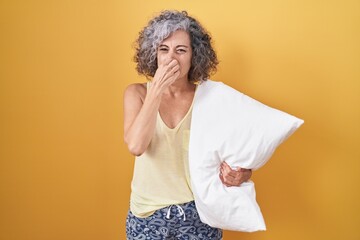 Middle age woman with grey hair wearing pijama hugging pillow smelling something stinky and...
