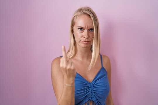 Young caucasian woman standing over pink background showing middle finger, impolite and rude fuck off expression