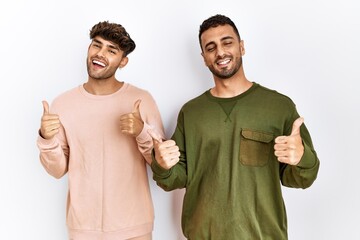 Young gay couple standing over isolated white background success sign doing positive gesture with hand, thumbs up smiling and happy. cheerful expression and winner gesture.