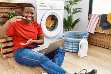 Young african man doing laundry and using computer gesturing with hands showing big and large size sign, measure symbol. smiling looking at the camera. measuring concept.