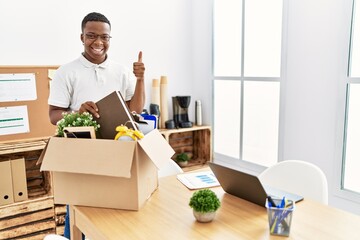 Fototapeta na wymiar Young african man putting office objects into cardboard box smiling happy and positive, thumb up doing excellent and approval sign