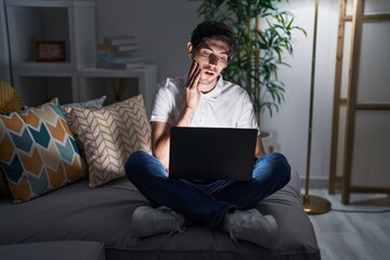 Young hispanic man using laptop at home at night touching mouth with hand with painful expression because of toothache or dental illness on teeth. dentist concept.