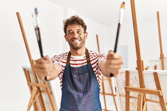 Young hispanic man smiling confident holding paintbrushes at art studio
