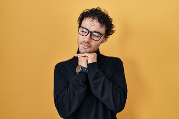 Hispanic man standing over yellow background laughing nervous and excited with hands on chin...