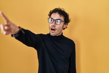 Hispanic man standing over yellow background pointing with finger surprised ahead, open mouth amazed expression, something on the front