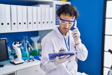 Young hispanic man scientist talking on smartphone reading document at laboratory